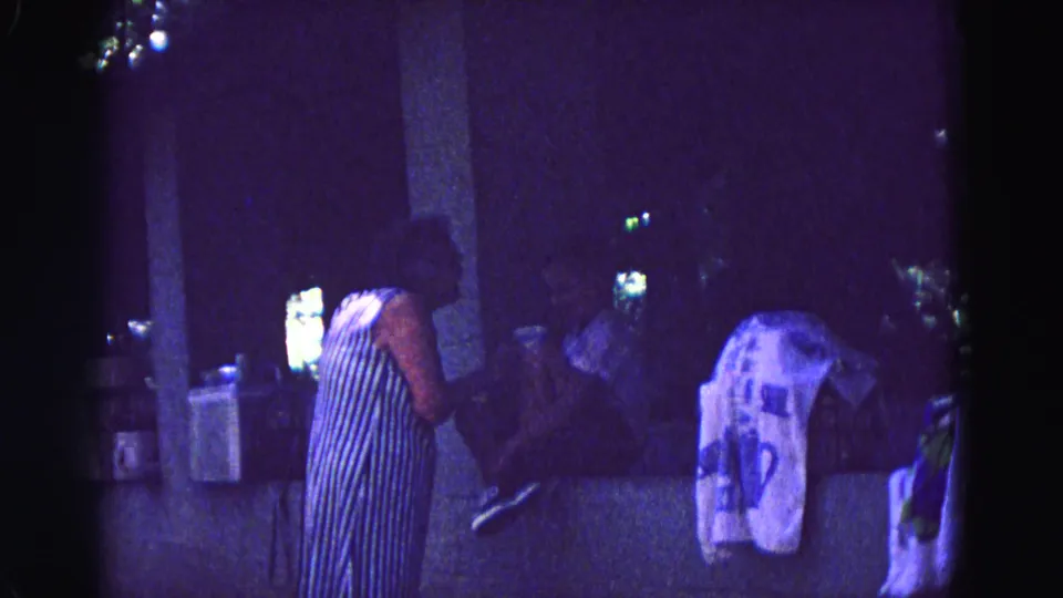 1967 Galena, Illinois home-movie frame with a standing woman, seated adults, pillars, and draped fabric in low purple-tinted light.