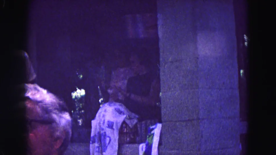 Purple-tinted 1967 Galena home-movie frame showing a foreground head, seated adults, a pillar, and draped cloth in a nighttime gathering.