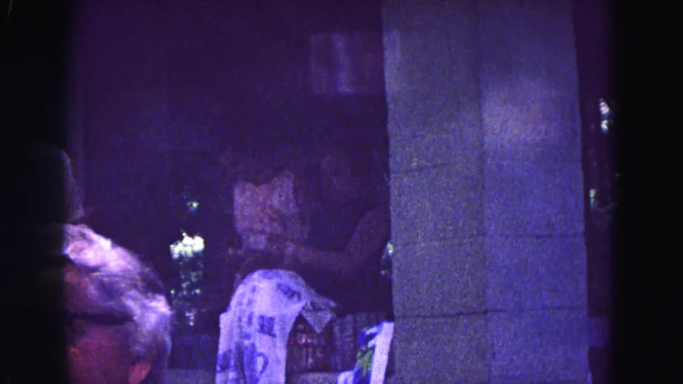 1967 Galena, Illinois 8mm frame with a foreground adult face, seated guests, a pillar, and draped fabric under very low light.