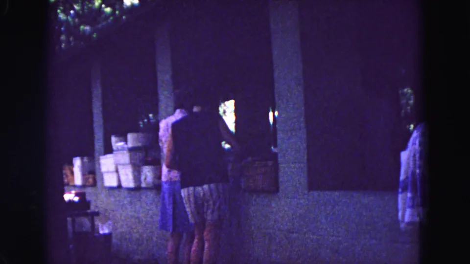 1967 Galena, Illinois home-movie frame showing women at a covered outdoor counter beside stacked boxes.