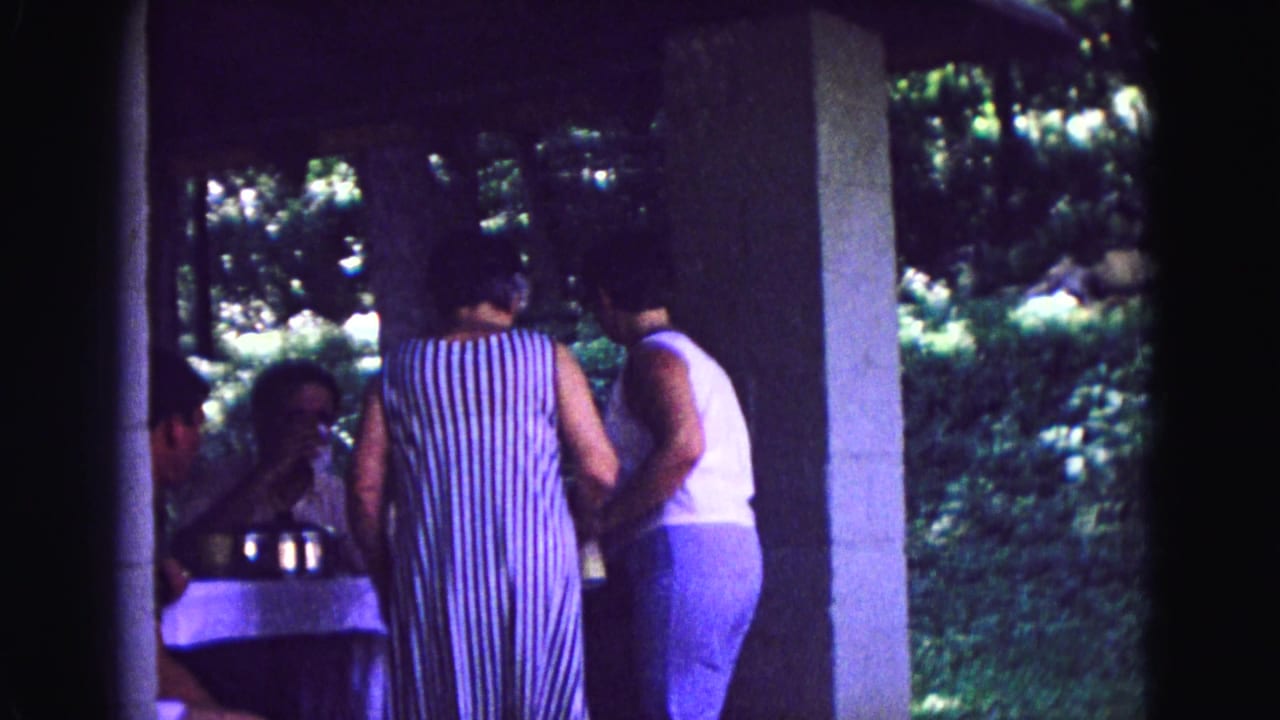 1967 Galena home-movie frame showing two women leaning near a patio table with a seated man under a covered outdoor area.