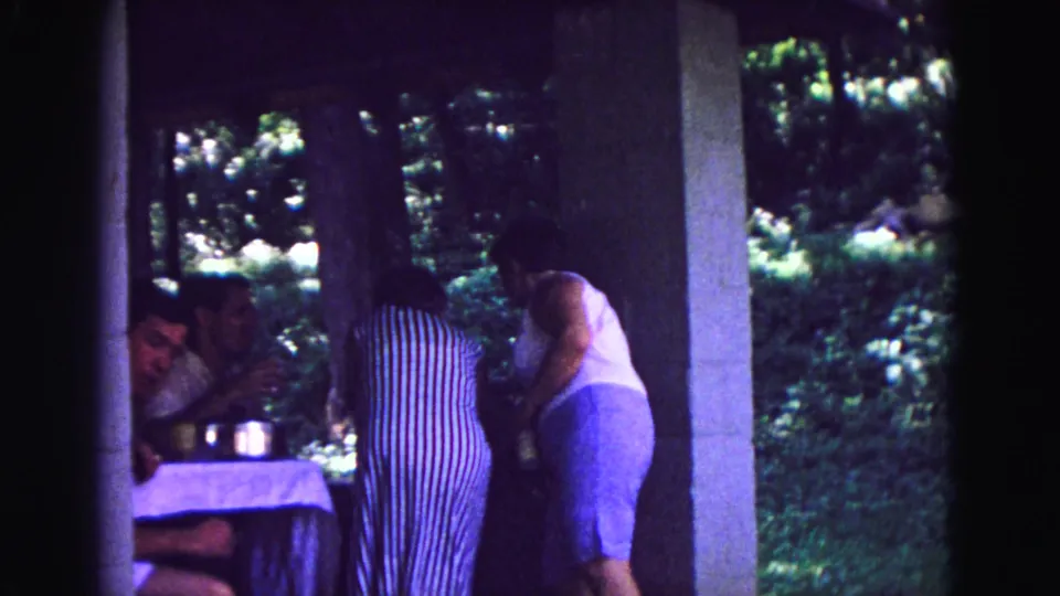 1967 Galena patio scene with two women leaning over table items while seated family members remain partly obscured in shade.
