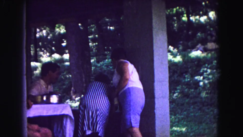Home-movie frame from Galena in 1967 showing women bending and standing beside a shaded patio table with greenery beyond.