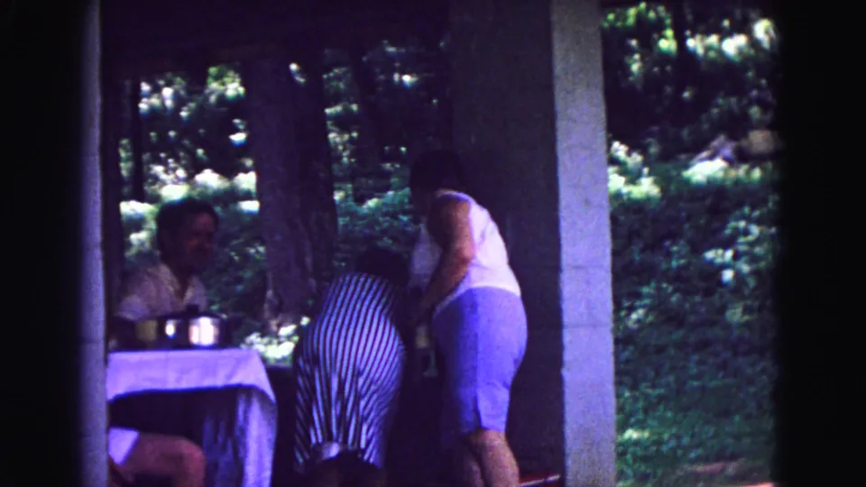 1967 Galena 8mm frame of a family patio moment, with women leaning near the table and a seated man watching from the shade.