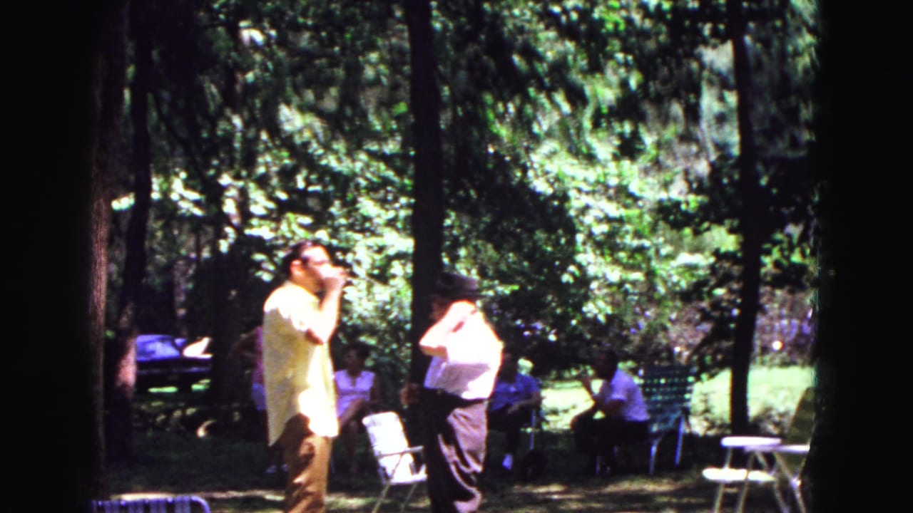 1967 Galena, Illinois frame with two men in light shirts standing in a sun-dappled wooded park while others sit behind them.