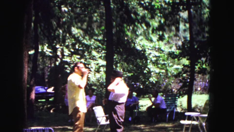 1967 Galena, Illinois frame with two men in light shirts standing in a sun-dappled wooded park while others sit behind them.
