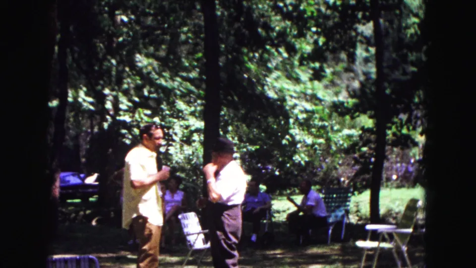 1967 Galena, Illinois home-movie frame showing two men talking near folding chairs with seated adults and dense trees behind them.