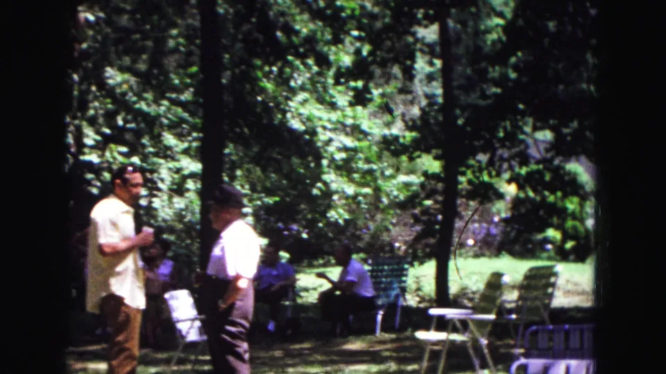 1967 Galena, Illinois frame of two men standing at a park picnic, with empty folding chairs and seated adults under trees.