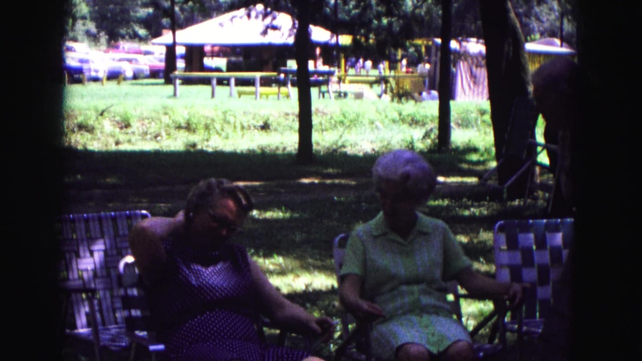 1967 Galena, Illinois home-movie frame showing two older women seated in shaded lawn chairs near a picnic shelter.