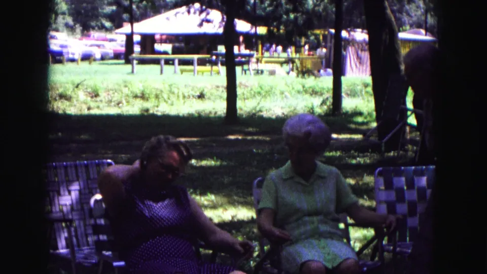 1967 Galena, Illinois home-movie frame showing two older women seated in shaded lawn chairs near a picnic shelter.