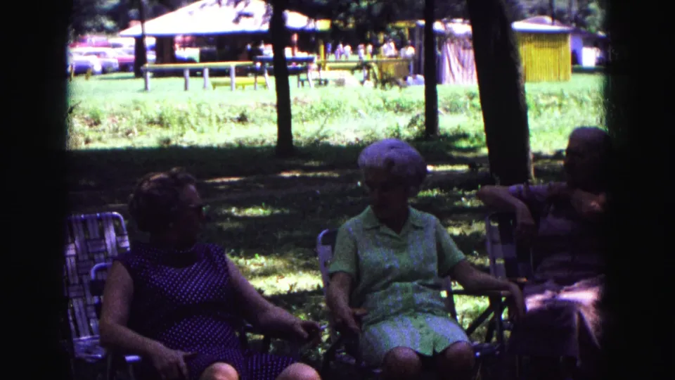 1967 Galena, Illinois frame showing older women in conversation on lawn chairs, with picnic tables and a yellow structure beyond.