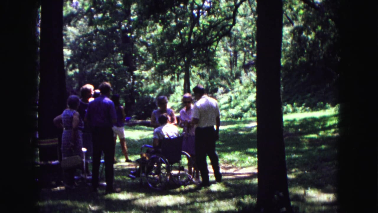 Group of adults gathered in a wooded area around a man seated in a wheelchair.
