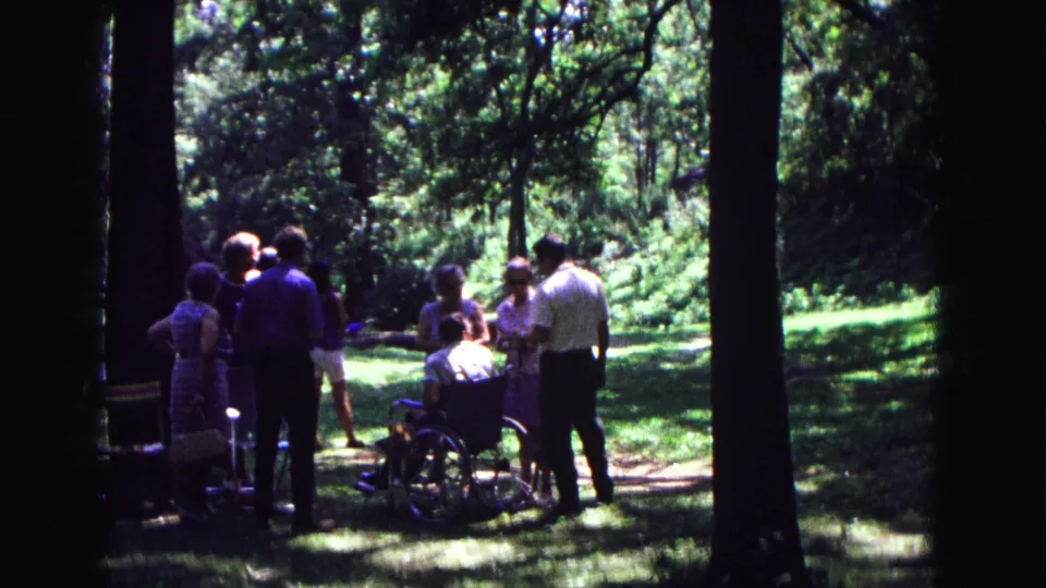 1967 Galena, Illinois home-movie frame showing adults standing in shade around a man in a wheelchair among trees and grass.