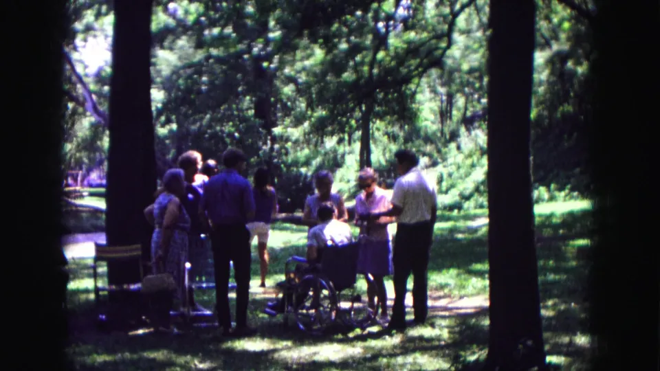 1967 8mm frame showing an informal outdoor gathering in Galena, Illinois, with adults around a seated wheelchair user.