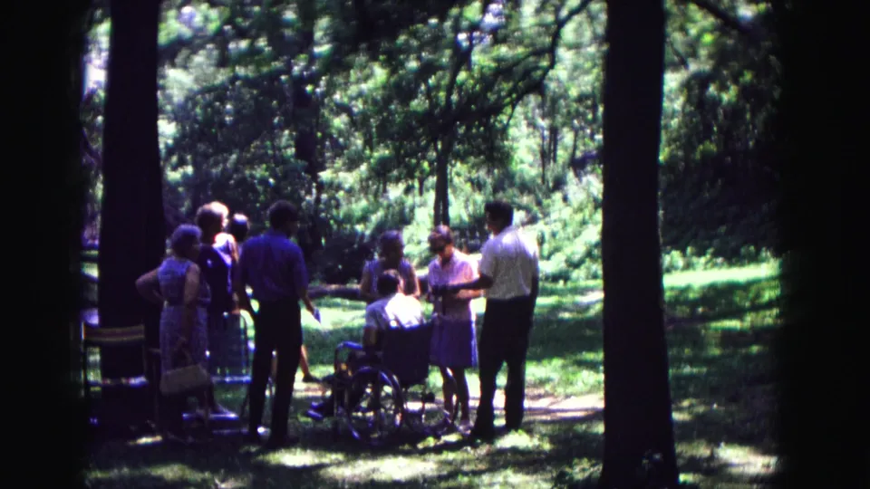 Late-1960s home-movie view of people talking outdoors in Galena, Illinois, framed by dark tree trunks and green lawn.