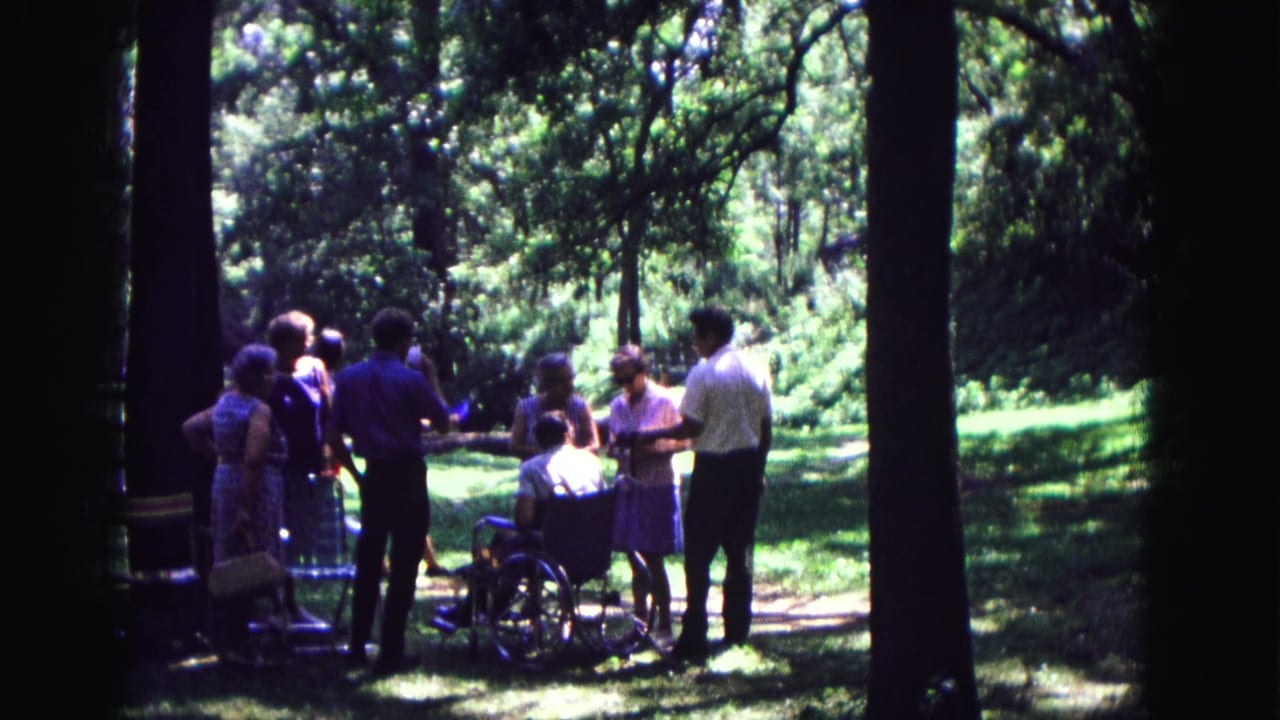 In Galena, Illinois, adults gather in a forest clearing around a man seated in a wheelchair, filmed on 8mm in 1967.