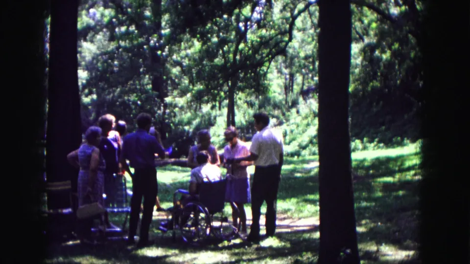 In Galena, Illinois, adults gather in a forest clearing around a man seated in a wheelchair, filmed on 8mm in 1967.