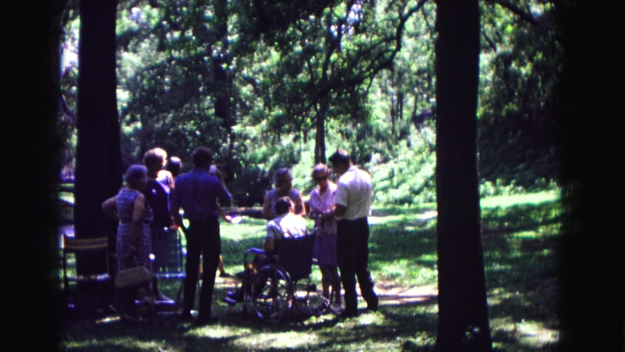 Group of adults converse around a wheelchair in a shaded forest clearing.