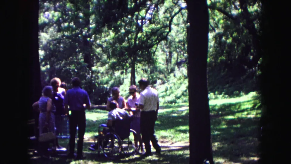 The 1967 8mm frame shows adults standing beside a wheelchair user in a wooded Galena clearing.