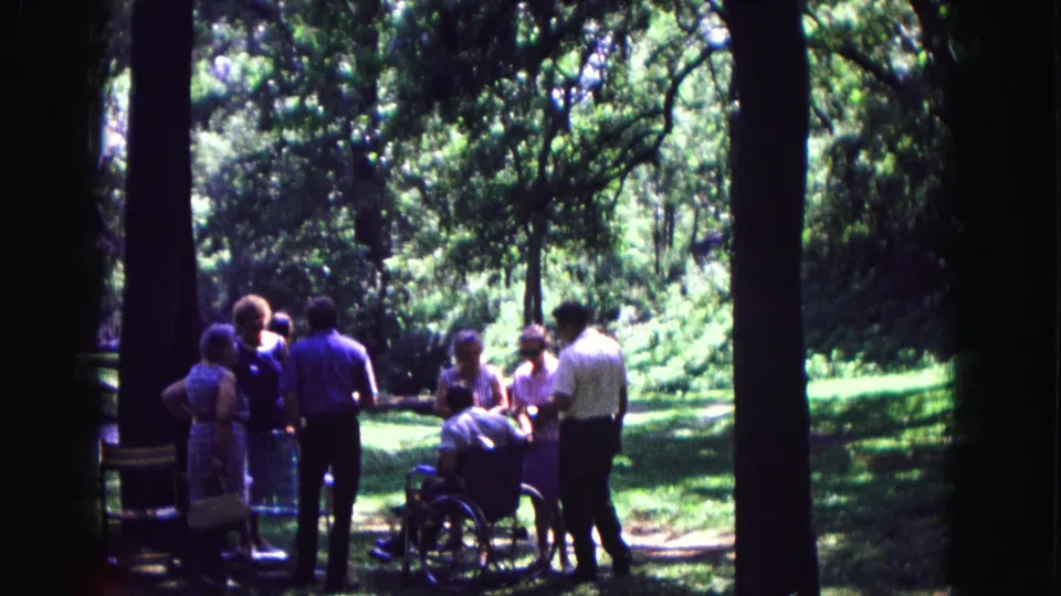 A small group in Galena, Illinois stands around a wheelchair user in dappled woodland light in 1967 home-movie footage.