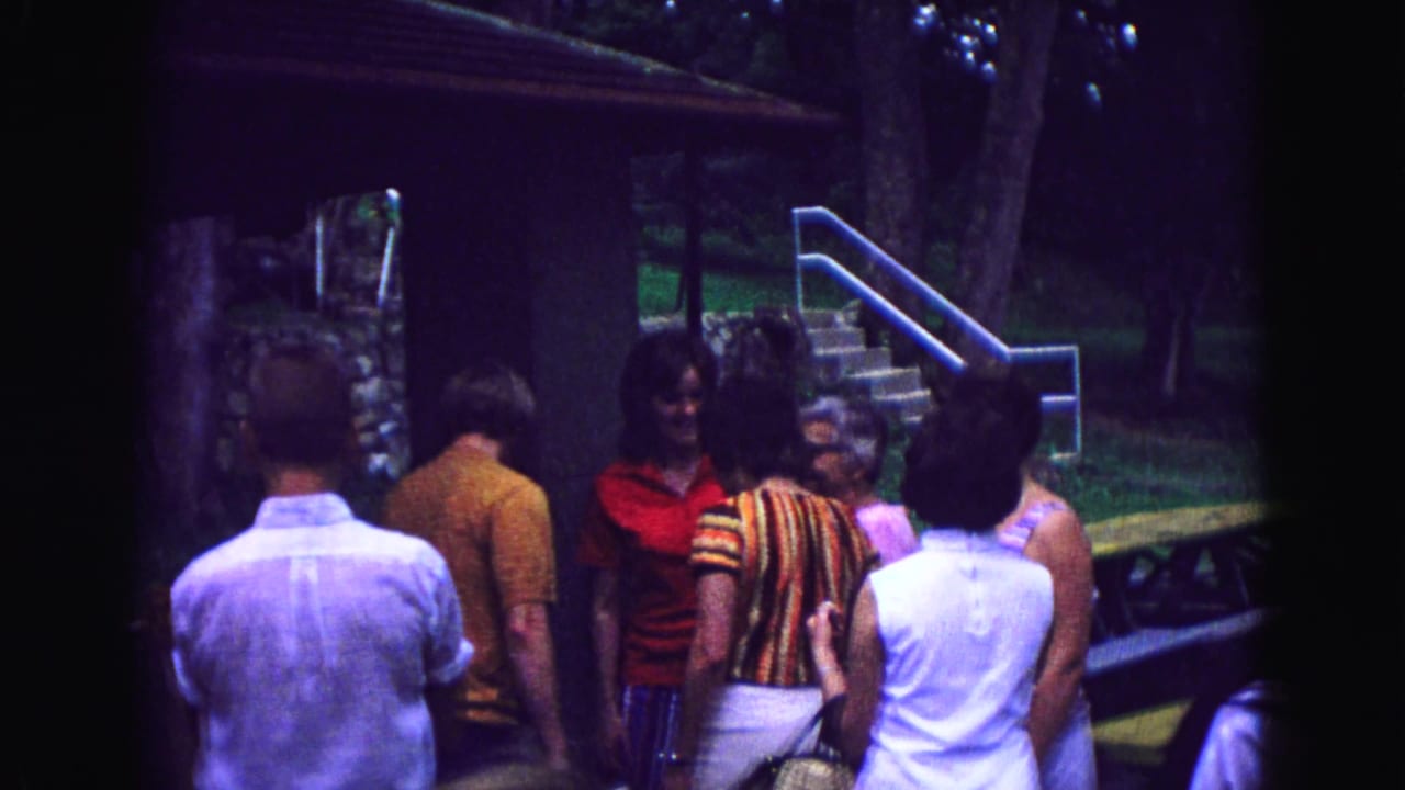 In 1967 Galena, Illinois home-movie footage, adults gather outside a rustic cabin near a stone wall, stairs, grass, and trees.