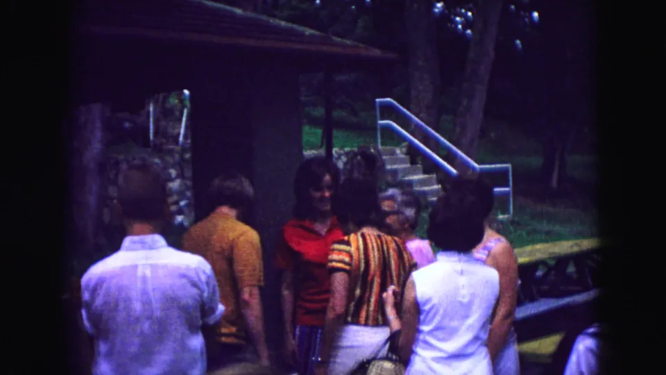 In 1967 Galena, Illinois home-movie footage, adults gather outside a rustic cabin near a stone wall, stairs, grass, and trees.