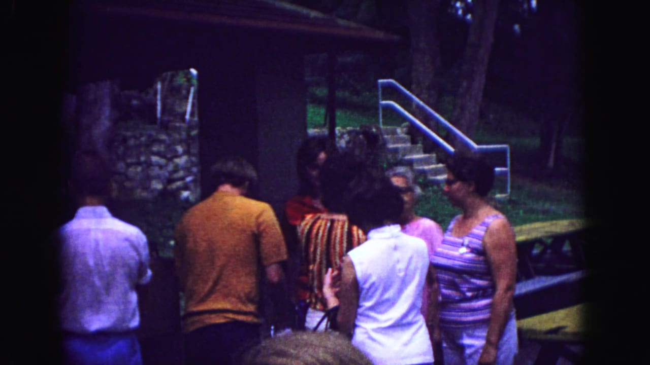 A 1967 Galena outdoor home-movie frame shows adults in casual clothing gathered by cabin stairs and a stone wall at dusk.