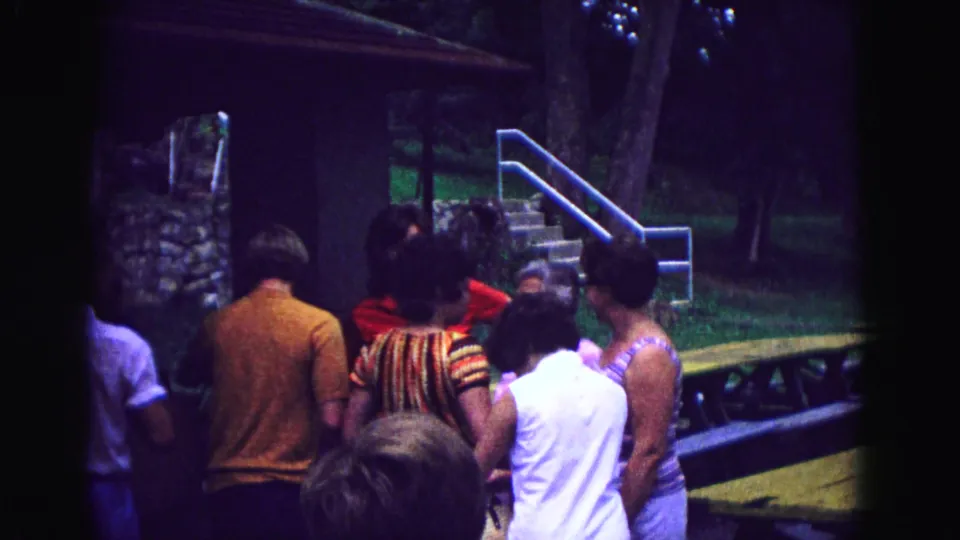 In the Galena, Illinois 1967 8mm footage, adults shift position while talking outside a rustic cabin near stairs and grass.