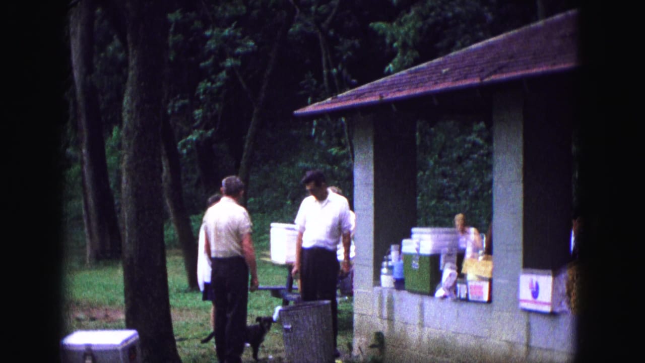 Two men stand near a picnic table and stacked coolers beside a park pavilion.
