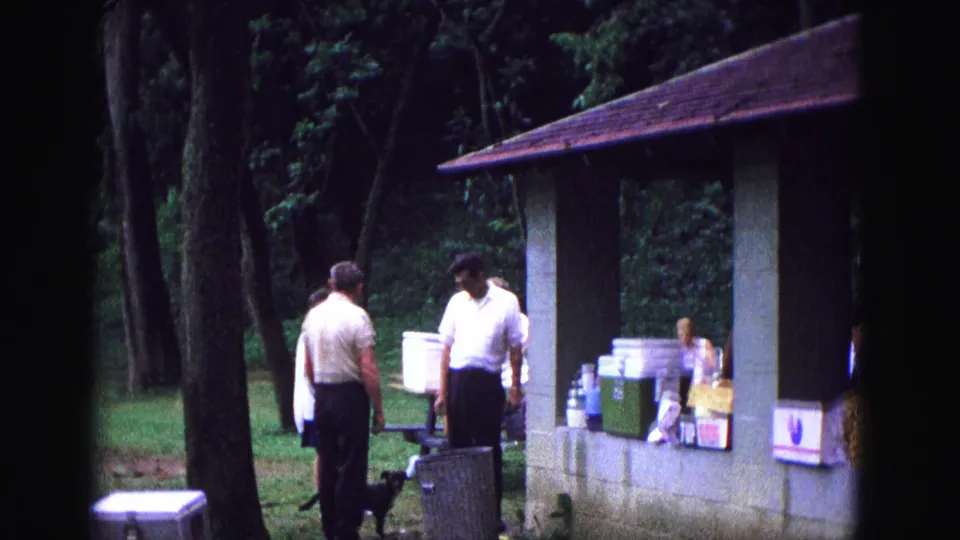 1967 Galena, Illinois picnic scene with two men, a dog, coolers, containers, trees, and a pavilion wall.