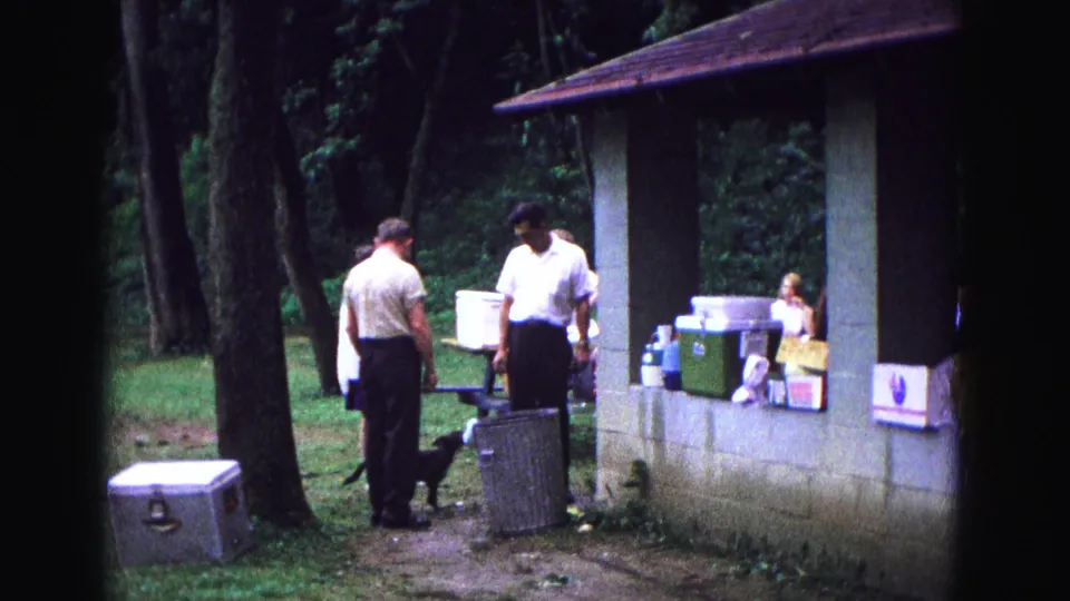 1967 Galena, Illinois home-movie still of men near picnic coolers, a dog, trees, and a concrete pavilion.