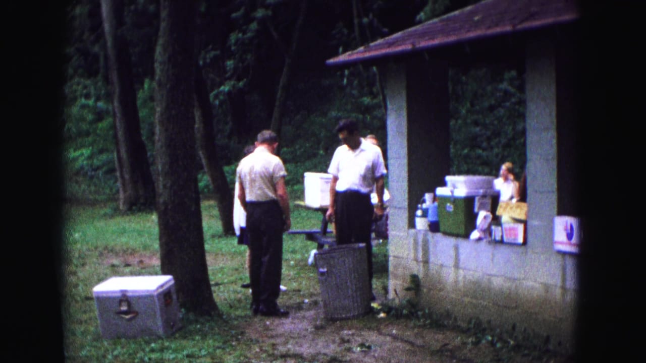 Adults standing near a concrete picnic shelter with coolers in a wooded park.