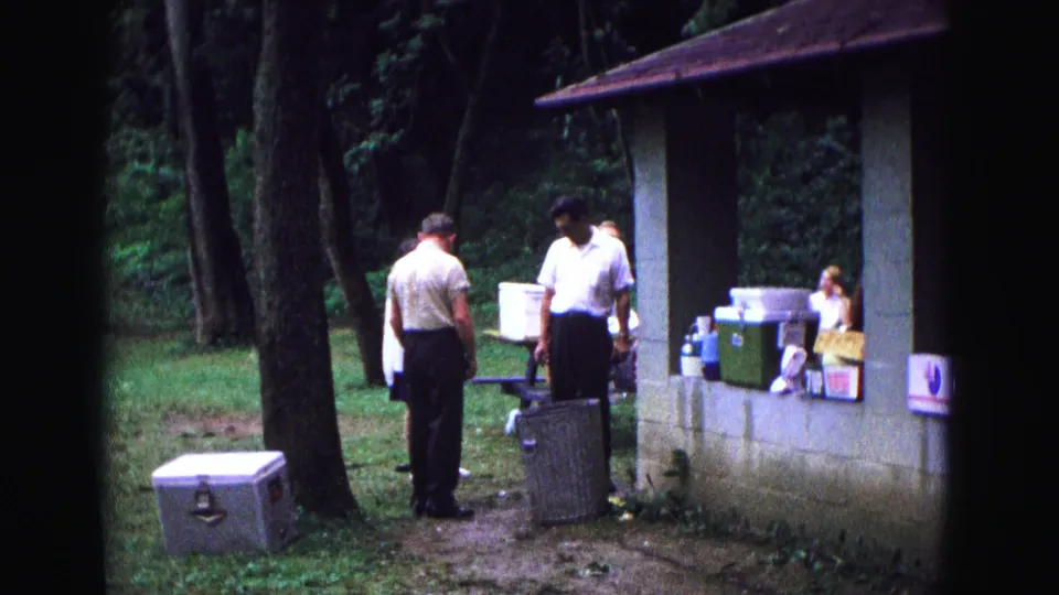 1967 home-movie frame from Galena, Illinois, showing adults near a concrete shelter, coolers, and picnic supplies among trees.