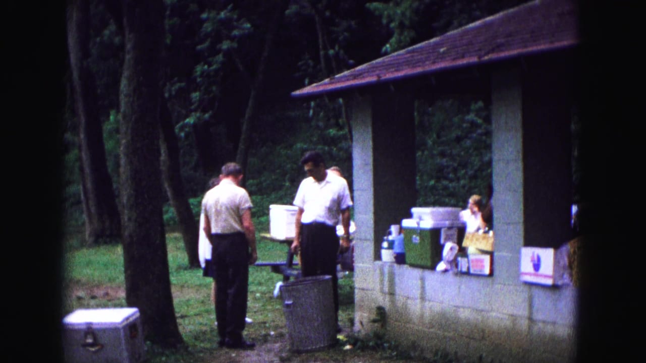 Men in short-sleeved shirts stand near coolers and food containers at a wooded Galena picnic shelter in 1967 home-movie footage.