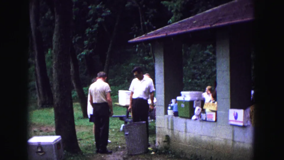 Men in short-sleeved shirts stand near coolers and food containers at a wooded Galena picnic shelter in 1967 home-movie footage.