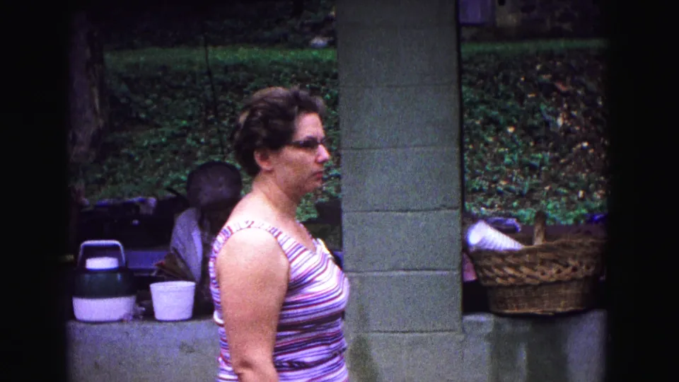 A 1967 home-movie frame shows the woman in profile at the picnic shelter, with cups, a cooler, and a wicker basket nearby.