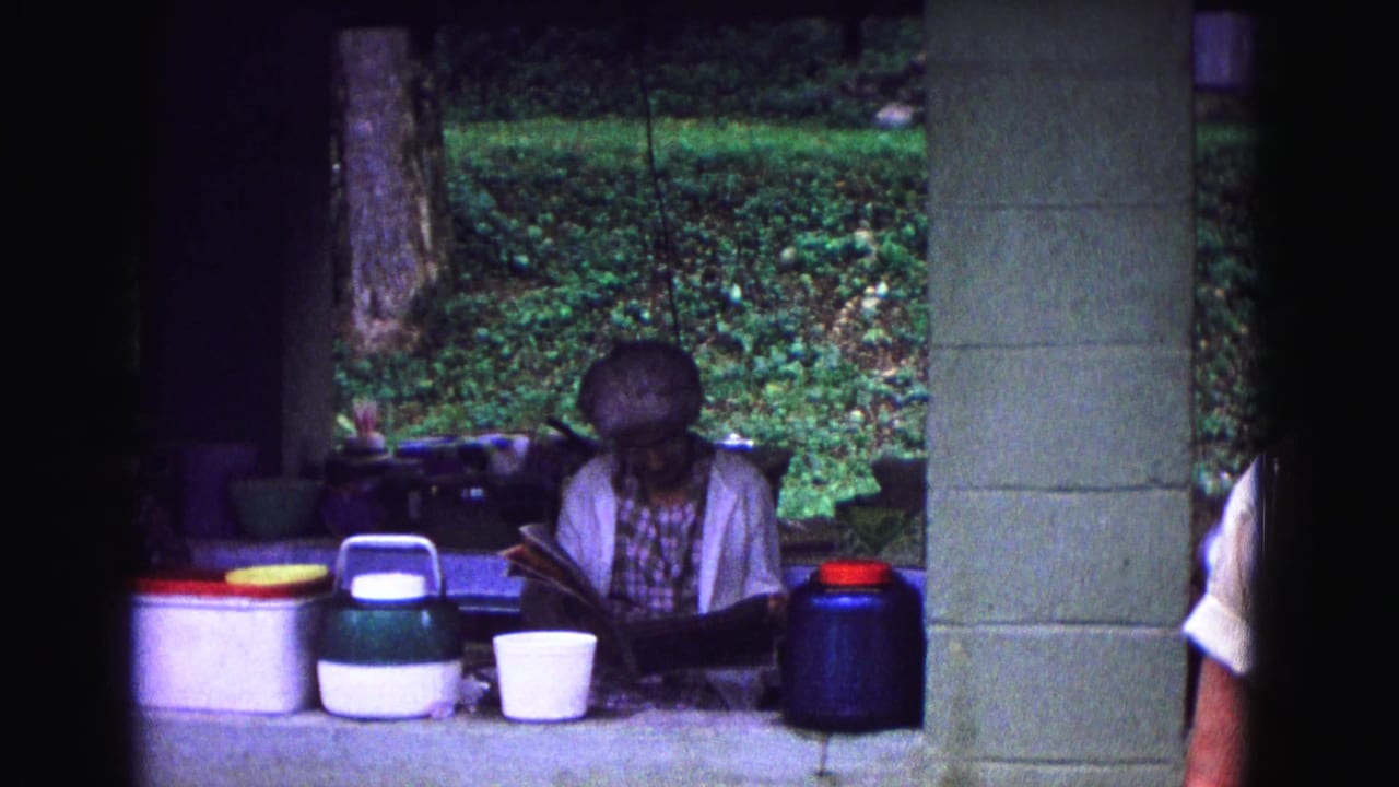In 1967 Galena, Illinois home-movie footage, a woman sits under a campsite shelter reading beside colorful food containers.