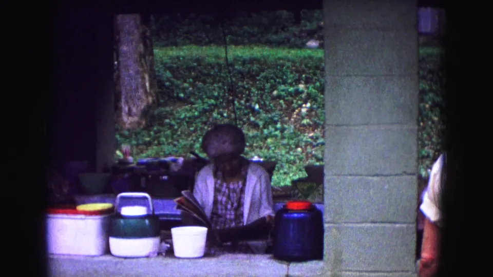 A closer 8mm frame shows the woman leaning over her reading material beside picnic containers at the Galena campsite shelter.