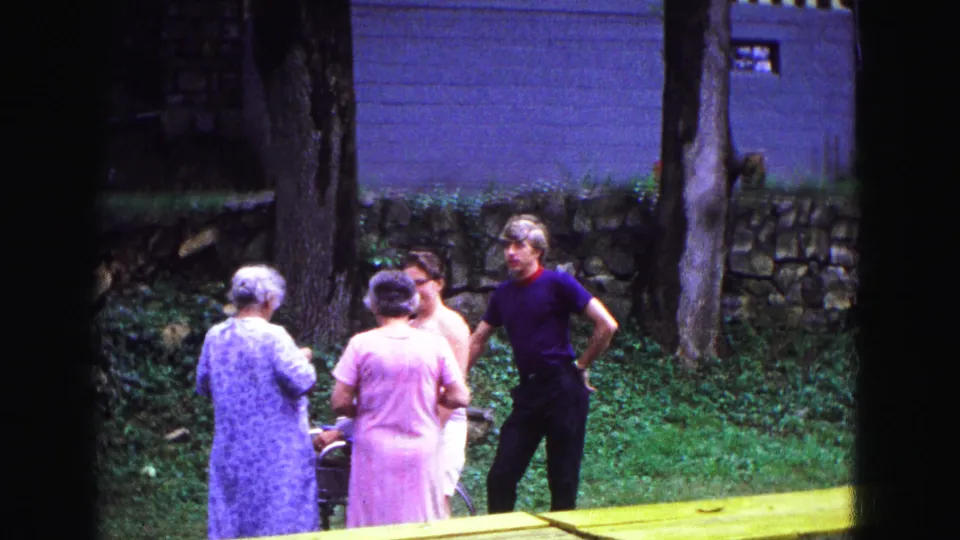 Several adults stand and talk in a grassy Galena yard near trees, a stone wall, and a building wall in 1967 home-movie footage.