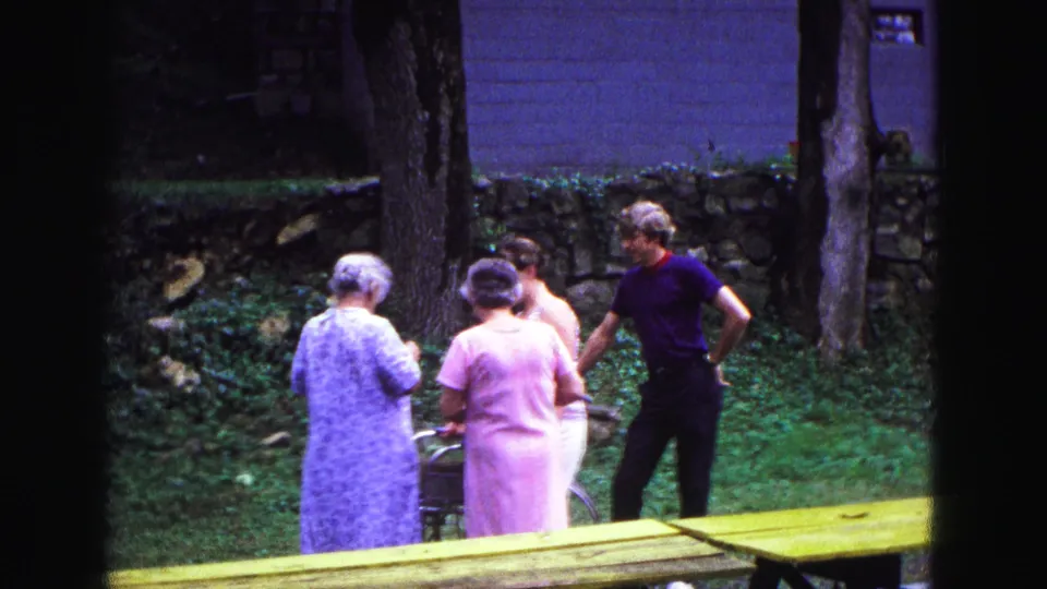 The Galena home-movie frame shows adults still gathered in a grassy yard, with a walker, tree trunks, and stone wall behind them.