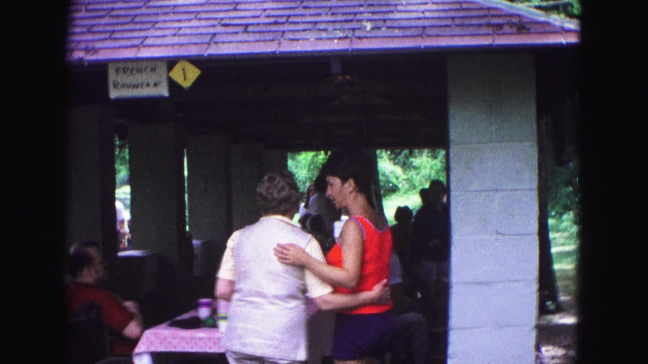 Two women greeting under a park shelter beside picnic tables and a small sign.
