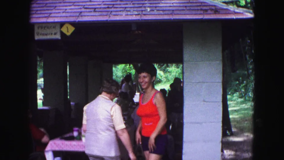 A woman in a red top smiles toward the camera near another woman beneath a Galena park shelter, with trees visible beyond.