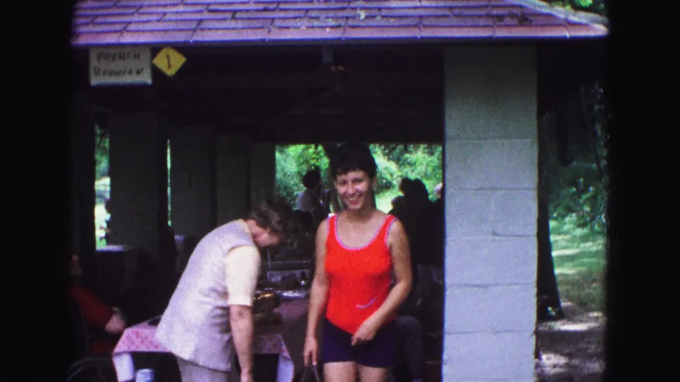 Under the park shelter, a woman in a red top faces the camera as another adult bends near a picnic table in the shaded gathering area.