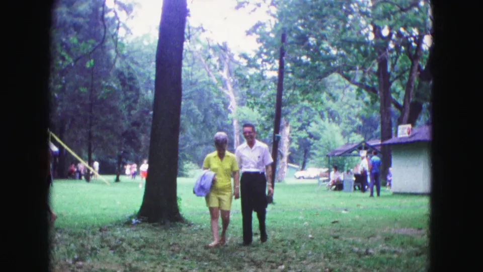 A man and woman walk across a tree-lined Galena park lawn in 1967 home-movie footage, with other visitors and a small building in the background.