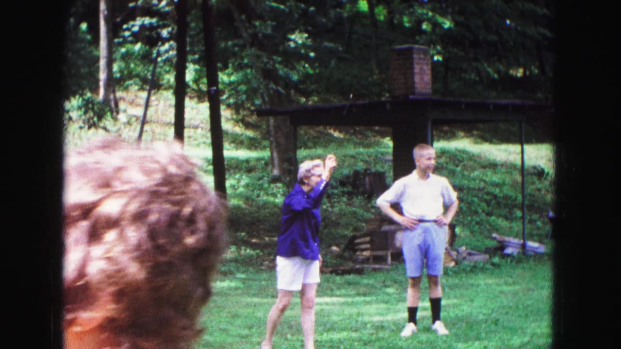 1967 Galena, Illinois home-movie frame showing adults on a wooded backyard lawn during a Frisbee game, with a shelter and brick chimney behind them.
