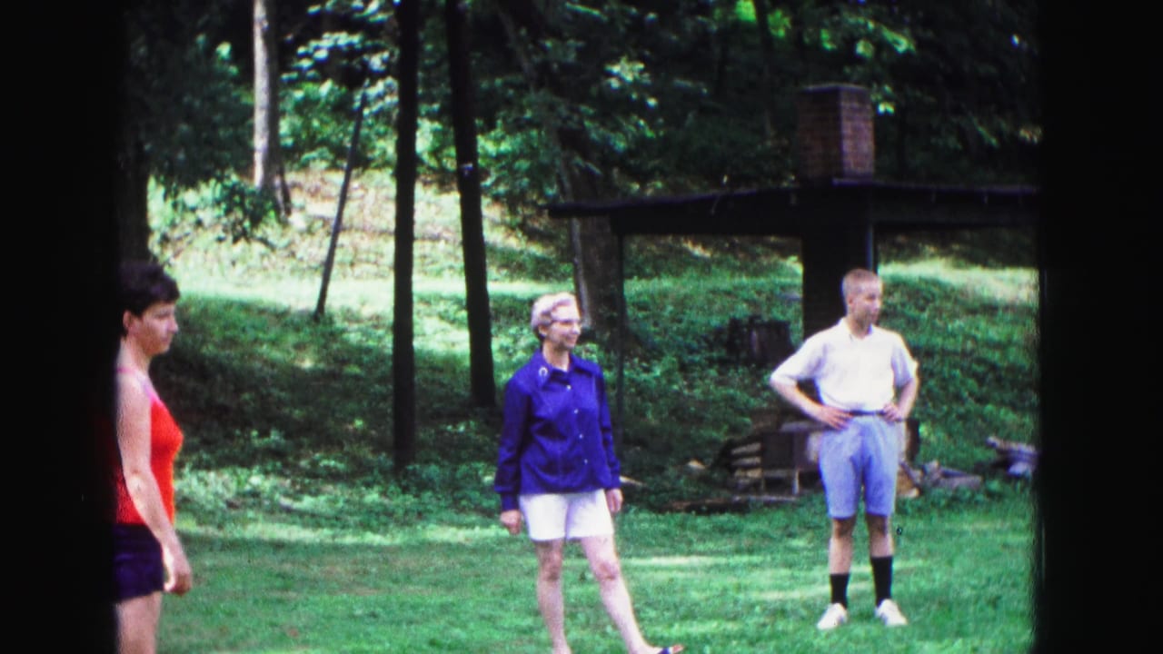 1967 Galena, Illinois frame with a woman in a red tank top at left and two adults waiting on a lawn near trees and a rustic shelter.