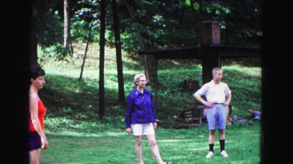 1967 Galena, Illinois frame with a woman in a red tank top at left and two adults waiting on a lawn near trees and a rustic shelter.