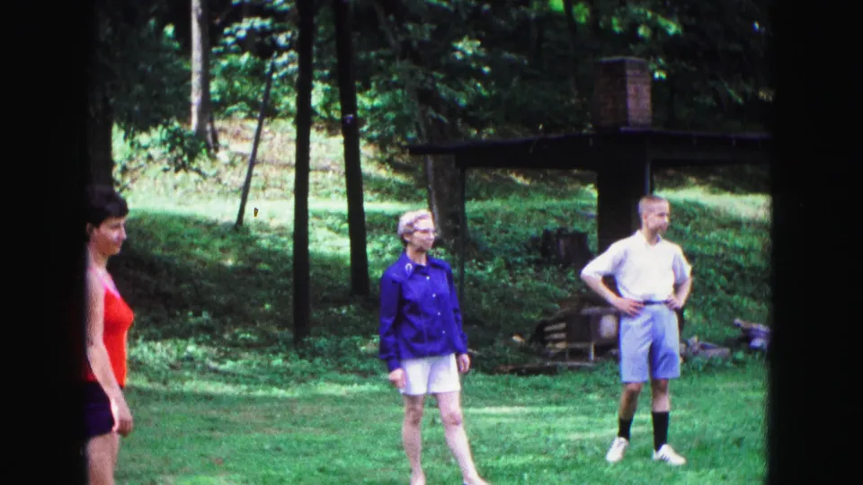 Home-movie frame from 1967 Galena, Illinois showing three adults in casual summer clothing on a wooded backyard lawn during Frisbee play.