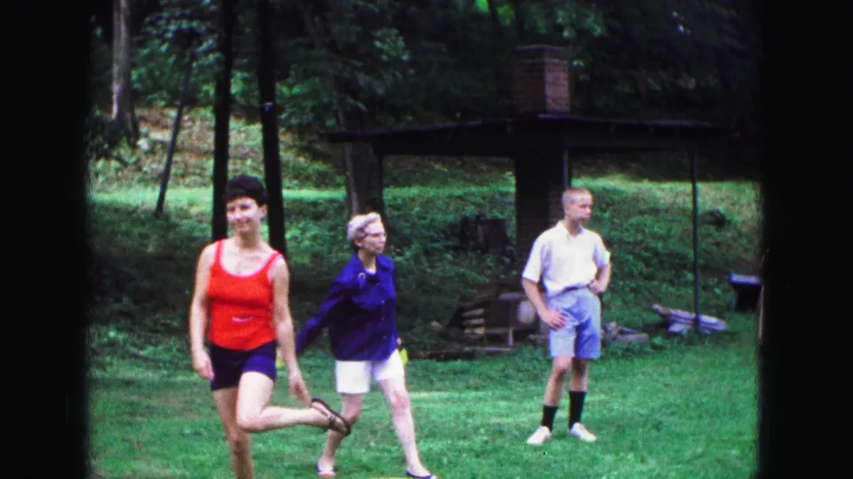 1967 Galena, Illinois home-movie frame showing a woman in red moving through a Frisbee game as two adults stand nearby on a wooded lawn.