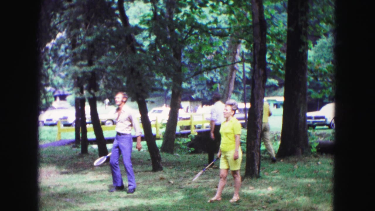 In Galena, Illinois in 1967, two adults stand with badminton rackets in a wooded park as period cars and a yellow fence sit behind them.